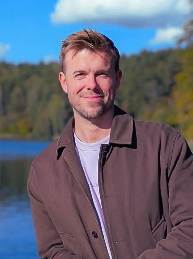 Alex smiling by a lake in a brown jacket, forest and blue sky behind